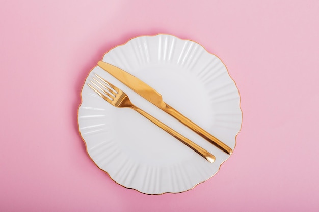 Beautiful white plate with golden knife and fork on pink background