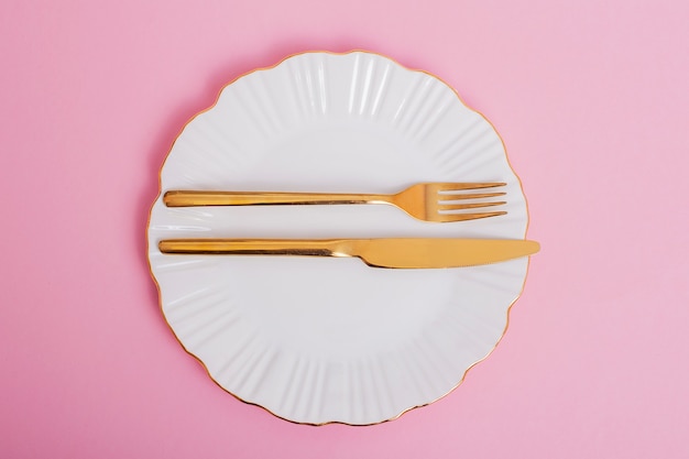 Beautiful white plate with golden knife and fork on pink background