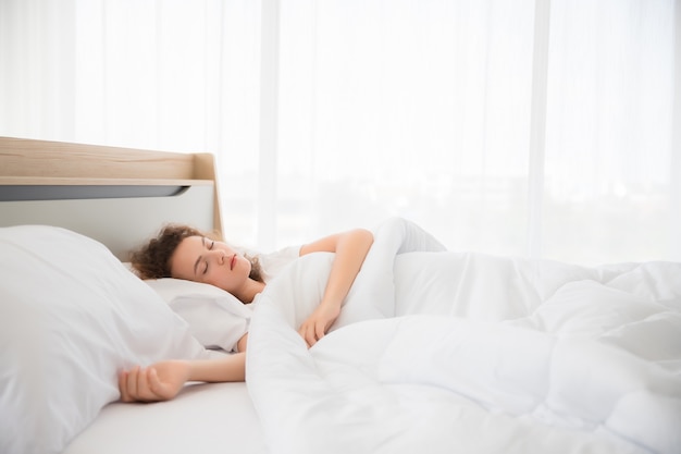 A beautiful white woman with curly hair is sleeping, resting and sleeping on a bed in a white bedroom.