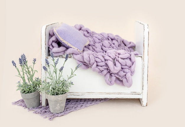Красивая белая деревянная кровать-мебель для студийной фотосессии новорожденного с украшением в фиолетовые цветы, подушкой и вязанным пледом. Крошечное место для детской фотографии на светло-розовом фоне