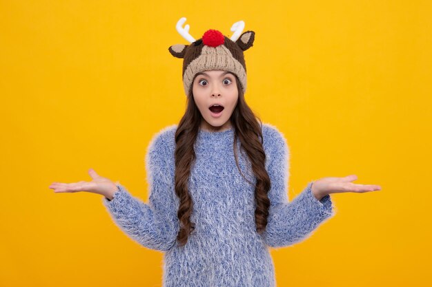 Beautiful winter kids portrait teenager girl posing with winter\
sweater and knit hat on yellow background surprised face surprise\
emotions of teenager girl