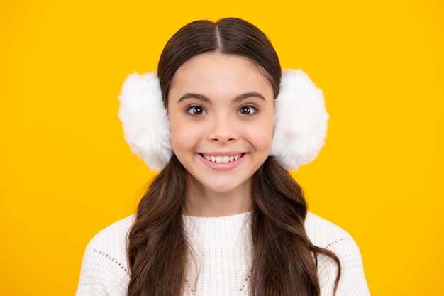 Beautiful winter kids portrait teenager girl posing with winter\
sweater and winter warm earmuff earflaps on yellow background happy\
face positive and smiling emotions of teenager girl
