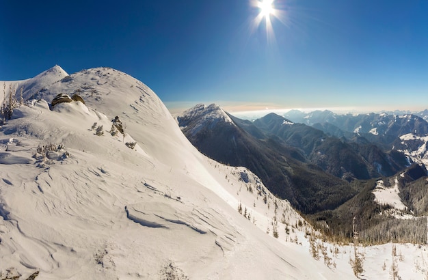 Beautiful winter landscape. Steep mountain hill slope with white deep snow, distant woody mountain range panorama stretching to horizon and bright shining sun rays on blue sky copy space