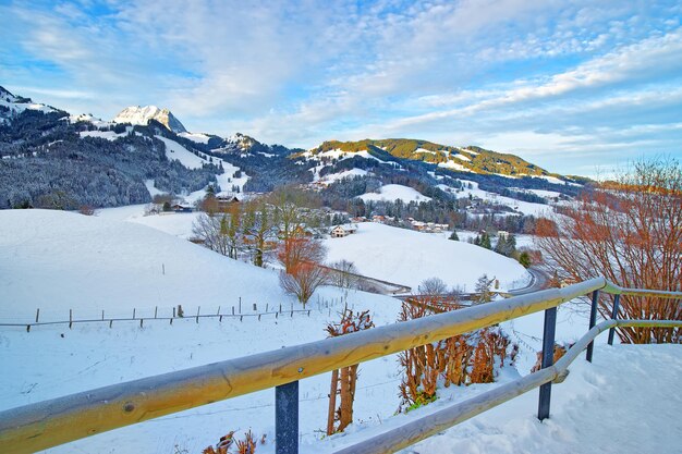 青い曇り空の下で雪に覆われた山の村と美しい冬の風景。スイス、フリブール州のグリュイエール