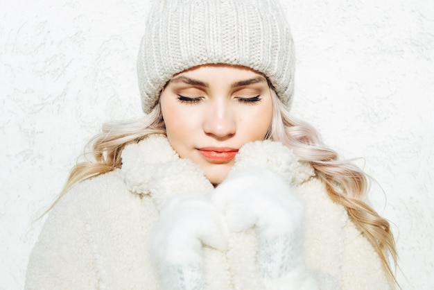 Beautiful winter portrait of young woman with eyes closed on white wall background.