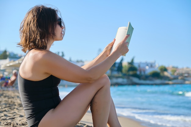 사진 40세의 아름다운 여성이 모래사장에서 휴식을 취하고, 책을 읽고, 바다와 일몰을 즐기고, 공간을 복사합니다.