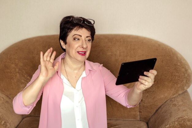 A beautiful woman of 60 years in glasses communicates with her relatives via video on a tablet.