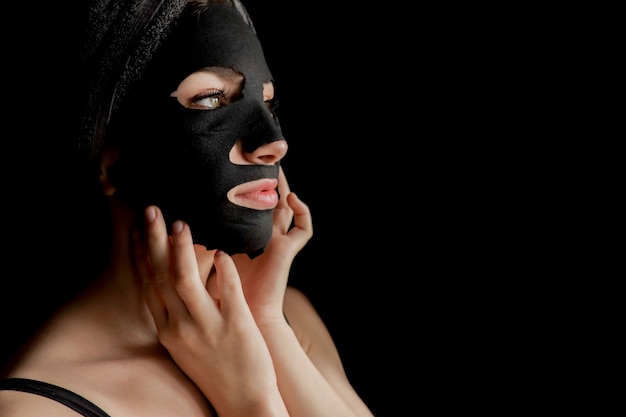 Bella donna che applica maschera facciale nera. trattamenti di bellezza. il ritratto del primo piano della ragazza della stazione termale applica la maschera di clay facial su fondo nero