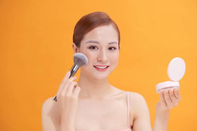 Beautiful woman applying blusher to her cheek with a large cosmetics brush while holding a compact mirror in front of her