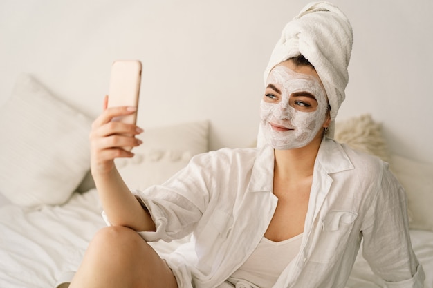 Beautiful woman applying facial mask and using mobile phone. 