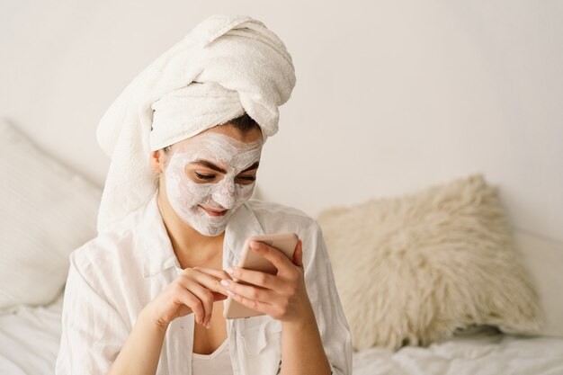 Beautiful woman applying facial mask and using mobile phone. 