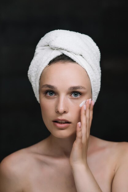 Beautiful woman applying moisturizing creme on face after shower Looking at camera Young pretty lady wrapped in towel smoothing perfecting skin daily morning routine concept