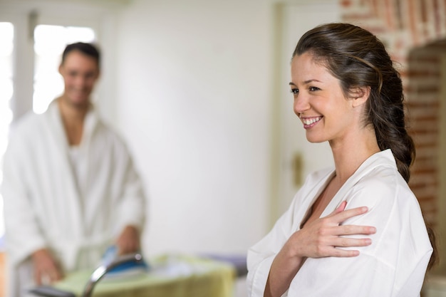 Beautiful woman in bathrobe looking away and smiling while man ironing clothes on background