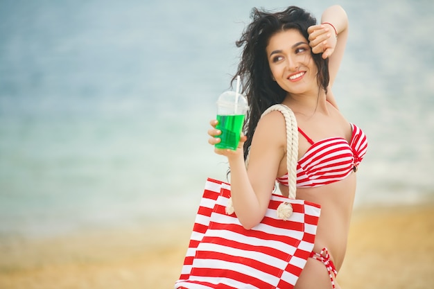 Beautiful woman at the beach having a tan in the red swimsuit with cold freshing drink and bag at the seaside in the summer heat