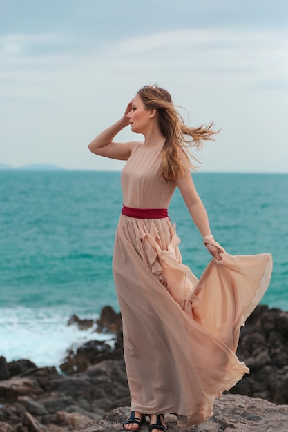 Beautiful woman in a beige dress stands on a rocky seashore