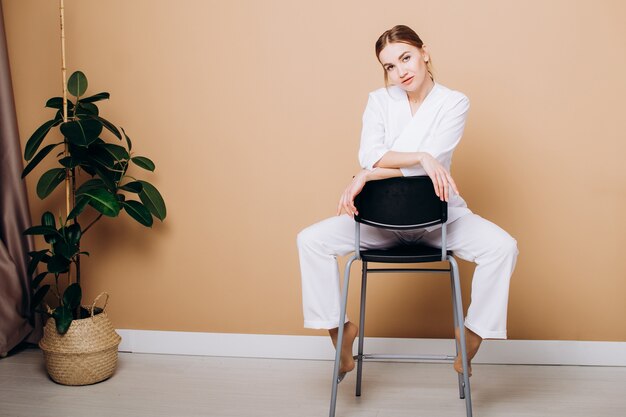 Beautiful woman in belote suit sits on a chair against the\
background of a brown wall