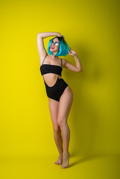 Beautiful woman in a black bikini in a colored wig and sunglasses on a yellow background girl posing in the studio