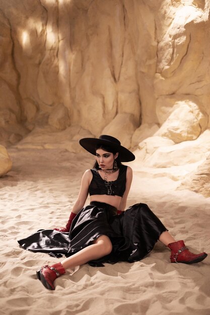 красивая женщина в черной шляпе и красных кожаных перчатках сидит на песке в каньоне, модный ковбой