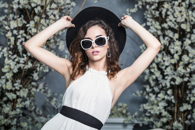 Beautiful woman in a black hat, sunglasses and a white dress against vintage white spring interior