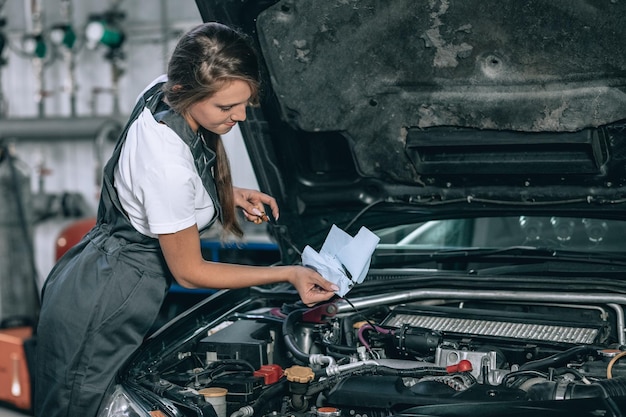 A beautiful woman in a black jumpsuit and a white t-shirt is smiling, checking the oil level in a black car in the garage.