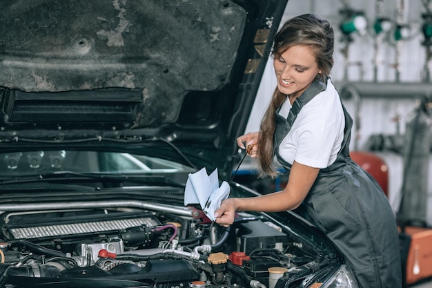 A beautiful woman in a black jumpsuit and a white t-shirt is smiling, checking the oil level in a black car in the garage.