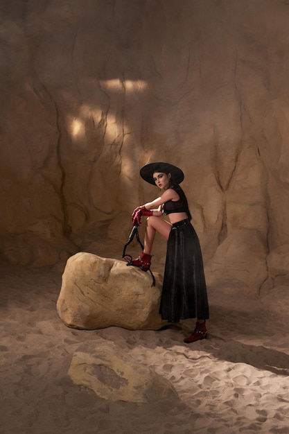 beautiful woman in black sexy clothes hat and red leather gloves with a whip near the rocks