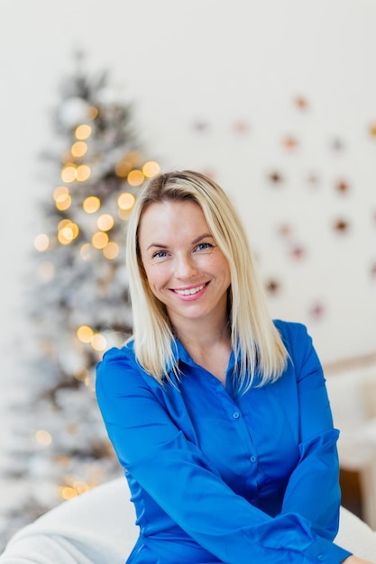 Beautiful woman in a blue dress sits on a white bed against the background of Christmas lights Girl smiling and looking at the camera