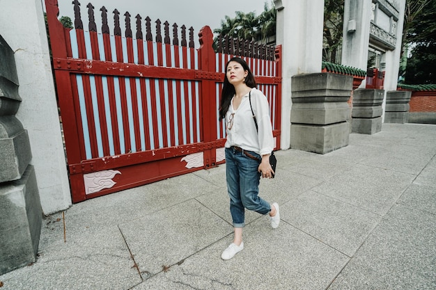 北京の中国の通りを一人で歩いているカジュアルな服装の美しい女性。若いアジアの日本人の女の子の観光客は歴史的記念物の観光を終えました。入り口の赤い門のそばの全身女性スタンド