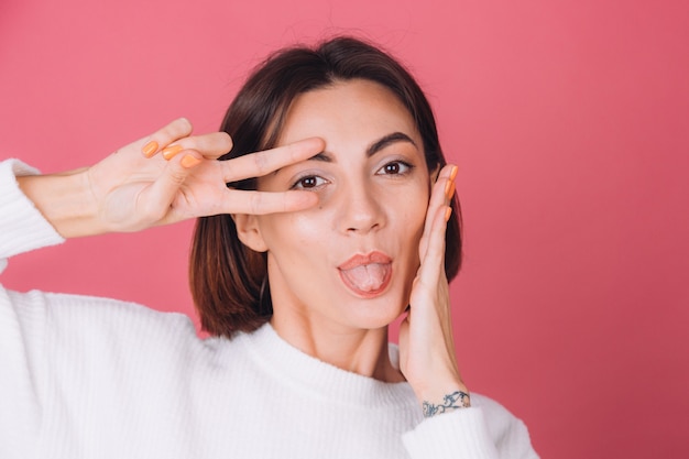 Beautiful woman in casual white sweater, does victory gesture shows tongue 