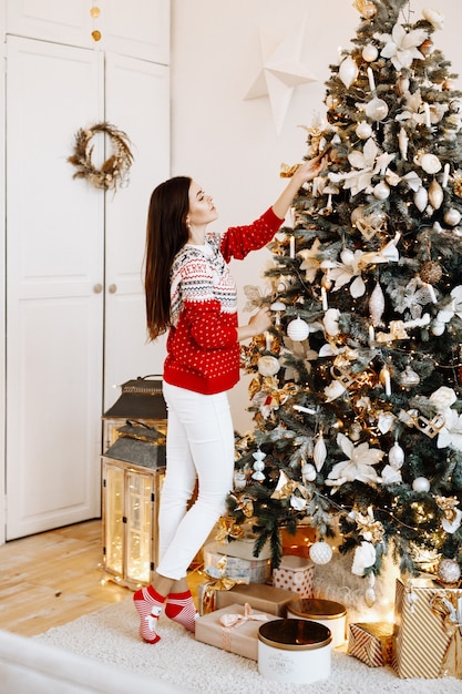 A beautiful woman in a Christmas sweater poses against a decorated fir tree