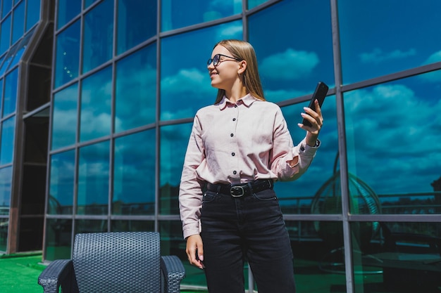 Beautiful woman in classic glasses and trendy outfit reading a message on a mobile phone standing next to a modern glass building