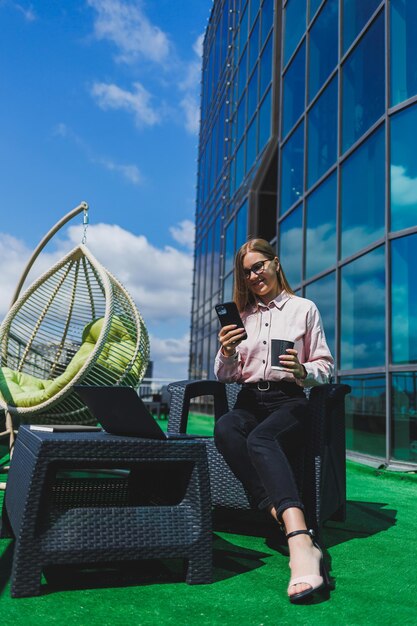 Beautiful woman in classic glasses and trendy outfit reading a message on a mobile phone standing next to a modern glass building