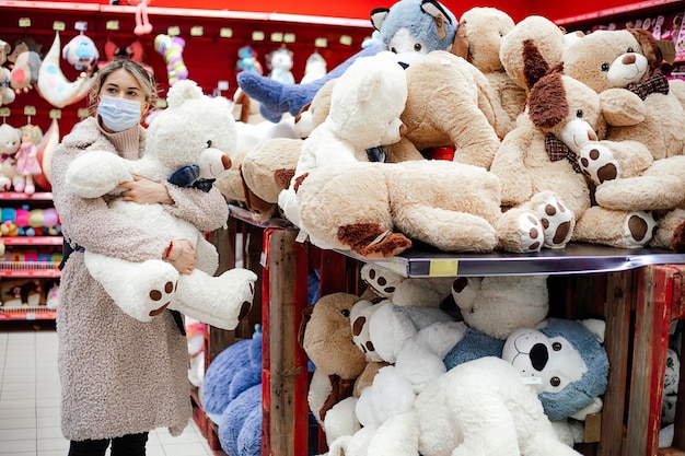 Beautiful woman in coat stands next to huge box of stuffed toys\
and holds large polar bear in her hands. children\'s toy store. new\
year\'s gifts. shopping during pandemic. protective equipment.