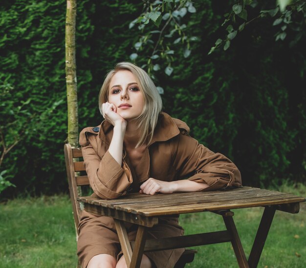 Beautiful woman in coat with blond hair sits at wooden table in a garden with trees on background