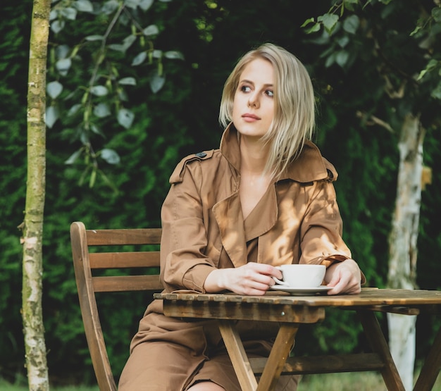 Beautiful woman in coat with cup of coffee at wooden table in a garden with trees on background