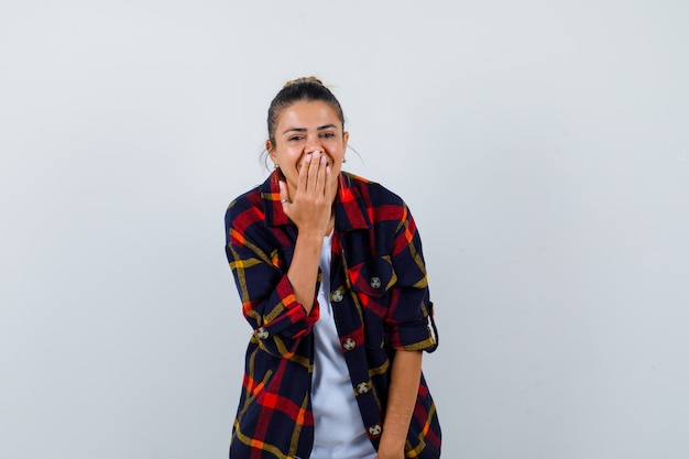 Beautiful woman covering mouth with hand, smiling in white t-shirt, cheked shirt and looking cheerful , front view.