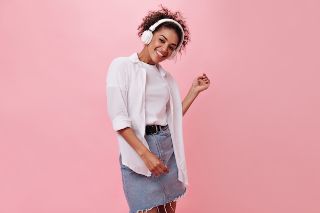 Beautiful woman in denim skirt listens to music with headphones on pink wall