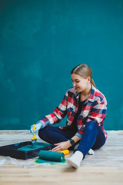 A beautiful woman designer makes repairs paints a gray wall with green paint with a brush Plaid shirt long hair and jeans Repair in the apartment