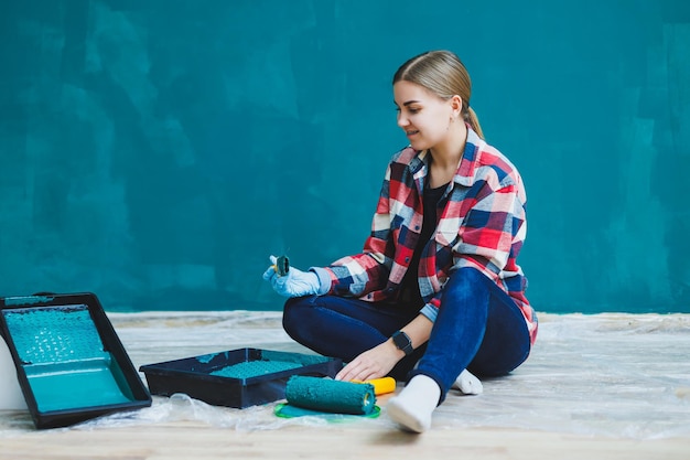 A beautiful woman designer makes repairs paints a gray wall\
with green paint with a brush plaid shirt long hair and jeans\
repair in the apartment