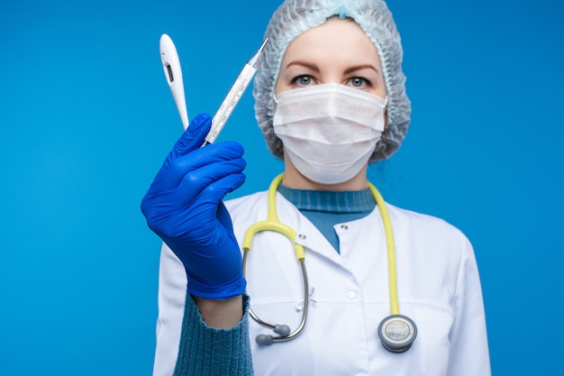 Красивая женщина-врач в белой медицинской одежде, маске, фонендоскоп и медицинской шляпе показывает электрические термометры, изображение, изолированных на синей стене