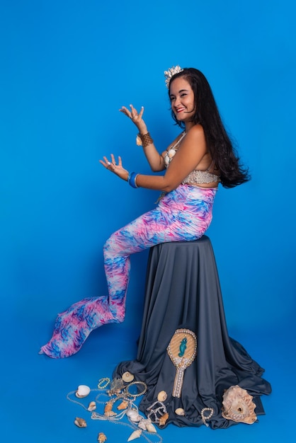 Photo beautiful woman dressed as a mermaid sitting looking at the camera next to several sea shells pearl necklaces and a mirror against a blue background