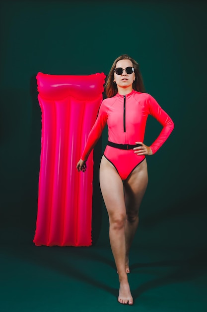 Beautiful woman dressed in pink swimsuit and sunglasses posing with pink inflatable mattress isolated on green background