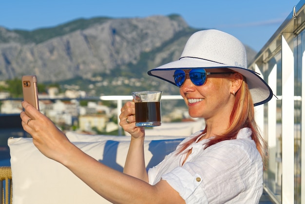 호텔 스파 리조트 야외 라운지 구역에서 밀짚 모자를 쓰고 신선한 커피를 마시는 아름다운 여성
