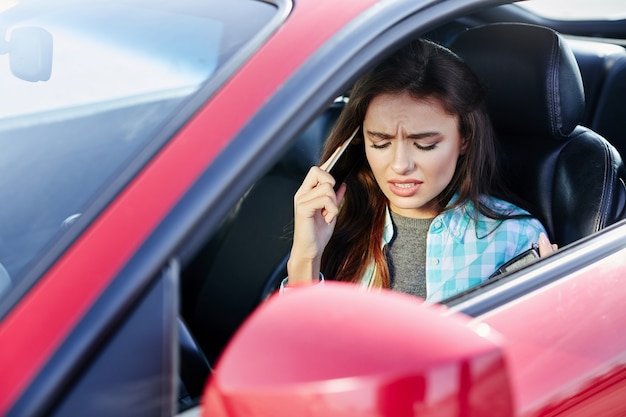 Beautiful woman driving red car, talk while driving. Profile of annoyed woman driving red car, stress while driving. Head and shoulders of brunette woman inside automobile