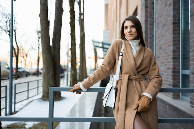 Beautiful woman in elegant brown coat and white bag posing\
outdoors