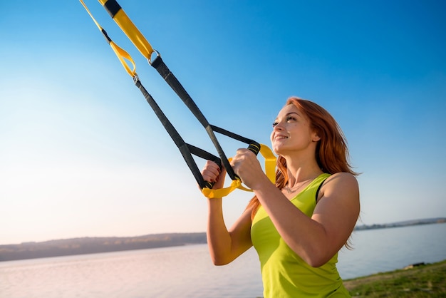 Красивая женщина тренируется с подвесными ремнями TRX на открытом воздухе возле озера в дневное время