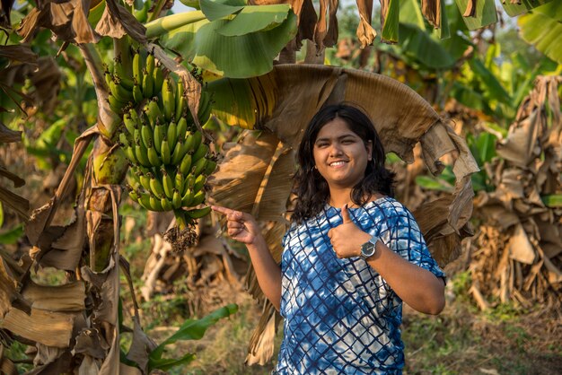 La bella lavoratrice agricola esamina o osservando o tenendo la frutta della banana sull'albero ad un'azienda agricola organica. sorriso volto di un contadino in anticipo nel campo dell'azienda agricola di agricoltura.