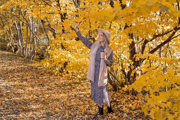 Beautiful woman in fashionable clothes and hat takes selfie on phone near yellow autumn tree