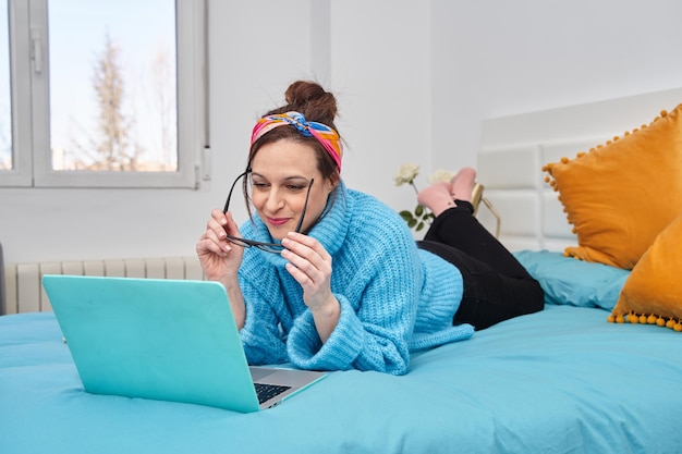 Beautiful woman in a fluffy blue sweater and glasses working from home on a laptop lying on the bed