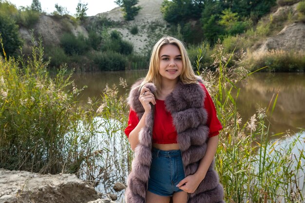 Beautiful woman in a fur coat and a light red T-shirt and shorts posing by the lake on a warm autumn day